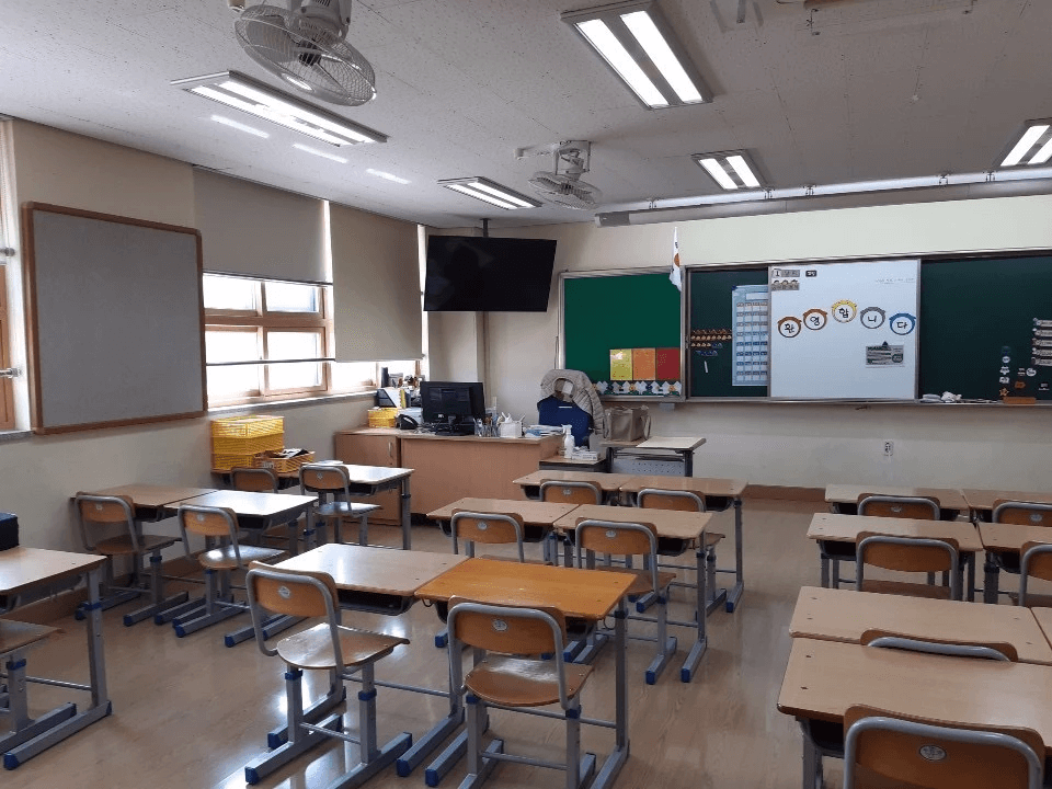 A typical elementary school classroom in Korea with a desktop on the teacher's desk with a big monitor above the desk on the wall for students to view.